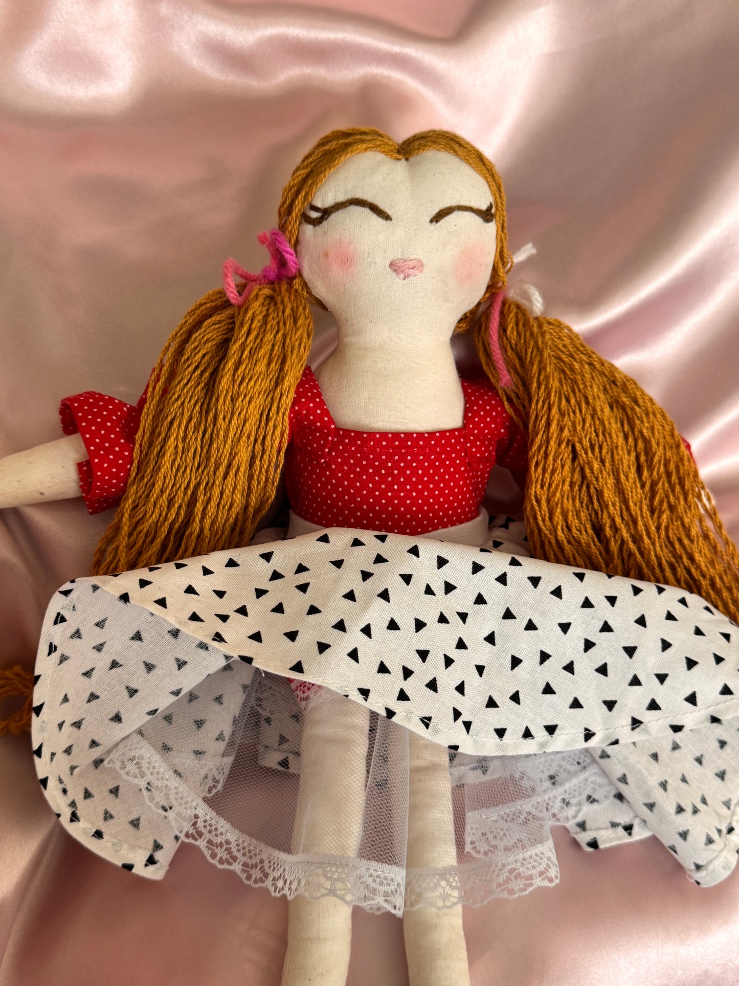 Textile doll with gold hair, red dress, and white lace dress on a pink fabric background