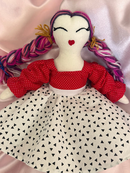Textile art piece with a doll-like figure wearing a red dress and holding a pink braided object, against a patterned background.